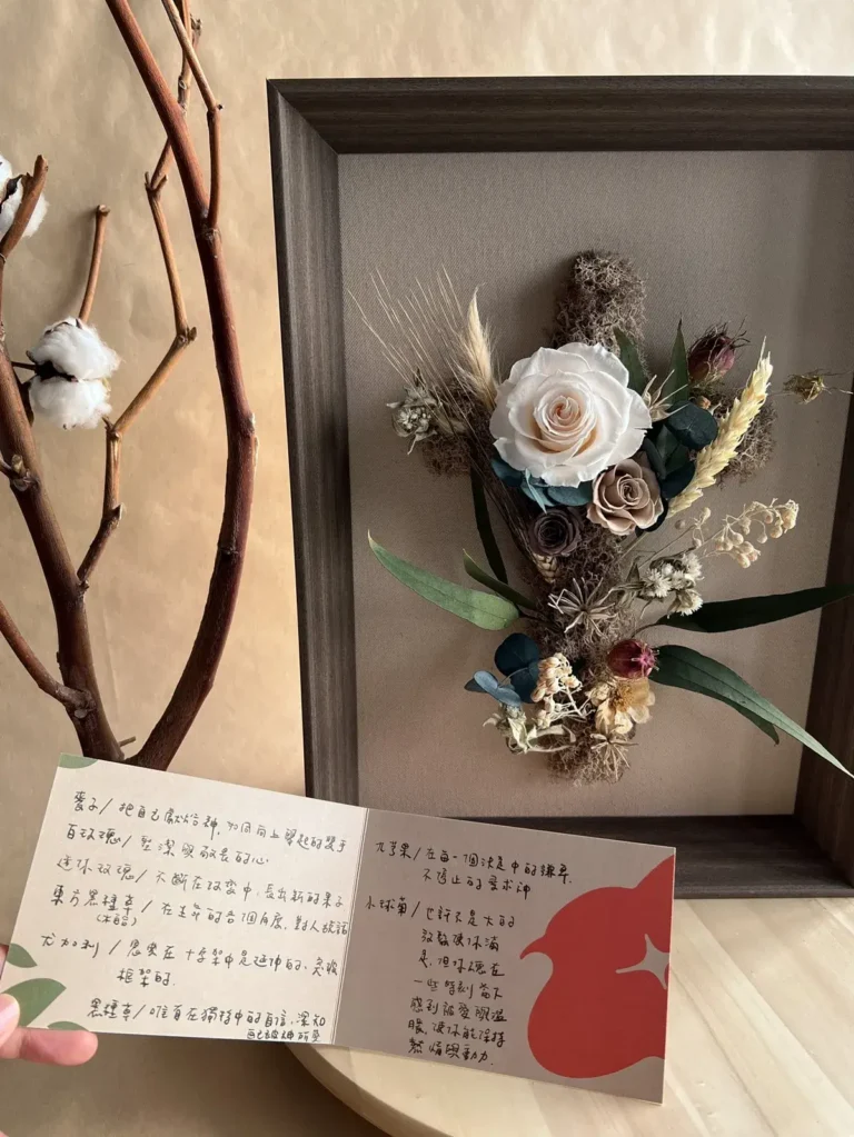 木框內的米白與大地色永生玫瑰十字架花藝，前方搭配手寫祝福卡片與棉花枝。